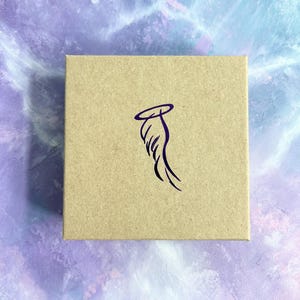 Pode incluir: Uma caixa de cart&atilde;o marrom com um design de asa de anjo roxo. A asa tem uma aur&eacute;ola acima.