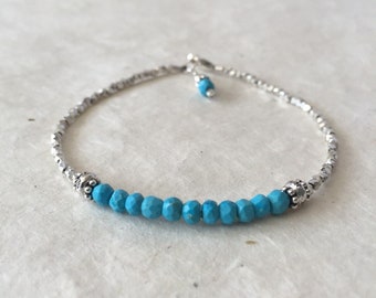 May include: A silver and turquoise beaded bracelet with a silver clasp. The bracelet is made with small, round beads and has a single row of turquoise beads in the center.