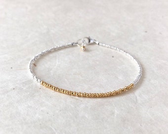 Puede incluir: Un brazalete delicado hecho con pequeñas cuentas plateadas y doradas. El brazalete tiene un diseño simple con una sola fila de cuentas.