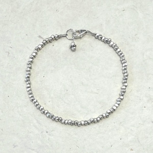 May include: A delicate bracelet composed of small, irregular, metallic silver-toned beads strung together in a circular pattern. The piece features a silver-toned lobster claw clasp and a small, textured dangling charm near the closure mechanism.