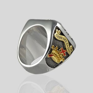 Puede incluir: Un anillo de plata con un dise&ntilde;o en negro y oro. El dise&ntilde;o presenta una corona, una cruz y una pancarta que dice "Cross and Crown".