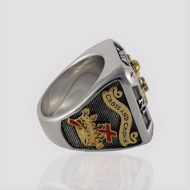 Puede incluir: Un anillo de plata con un dise&ntilde;o dorado y rojo. El anillo presenta una cruz y una corona con las palabras "Cross and Crown" inscritas en la banda.