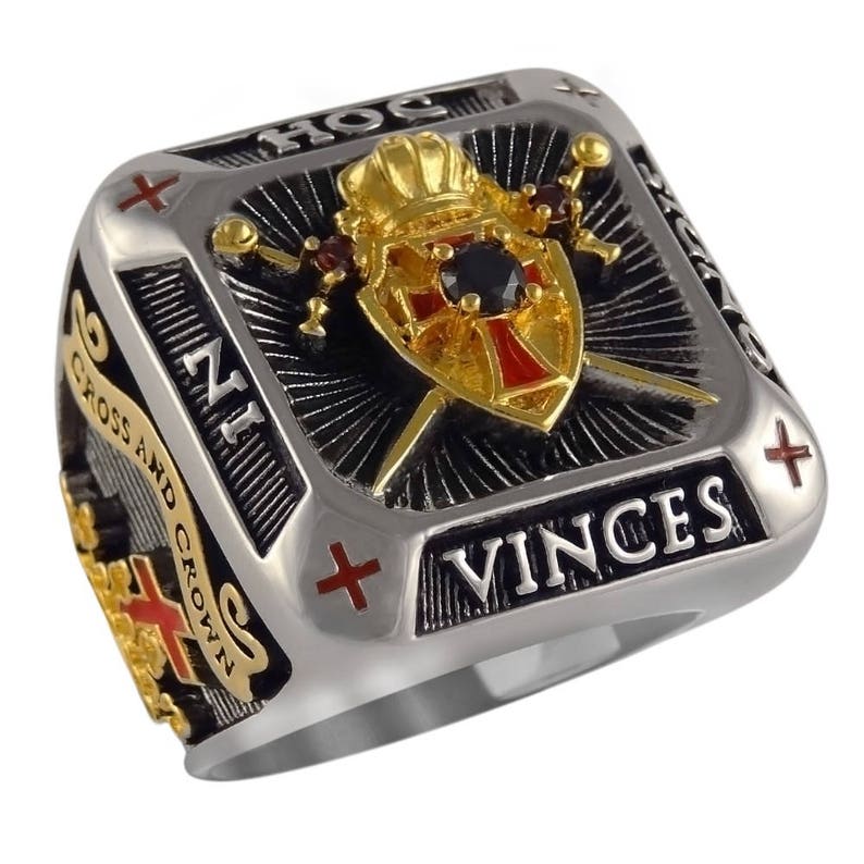 Puede incluir: Un anillo de plata y oro con fondo blanco y negro. El anillo presenta un escudo con una cruz roja, una corona y dos espadas. El texto "HOC VINCES" est&aacute; inscrito en el anillo.