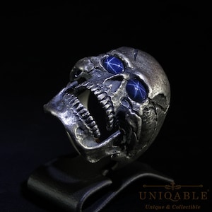 May include: A silver skull ring with open mouth and blue star-shaped gemstones for eyes. The ring has a detailed, textured surface and is displayed on a black stand. The ring is a unique and collectible item.