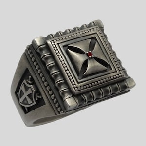 May include: A silver ring with a square design featuring a cross with a red stone in the center. The ring has a detailed design with a textured band.