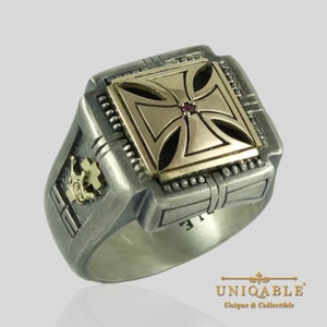 May include: A silver ring with a gold cross design and a red stone in the center. The ring has intricate details and a square shape.