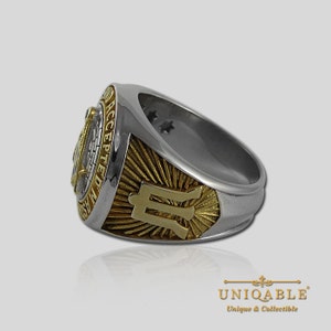 Pu&ograve; includere: Un anello in oro e argento con un simbolo massonico e le parole "In This We Trust Accepted" sul lato. L'anello ha una fascia in oro con un design a raggiera e la lettera "M" al centro.