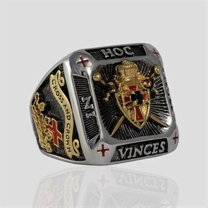 Puede incluir: Un anillo de plata y oro con un dise&ntilde;o de esmalte negro y rojo. El anillo presenta un escudo con una cruz, una corona y dos espadas. El texto "HOC VINCES" y "CROSS AND CROWN" est&aacute; inscrito en el anillo.