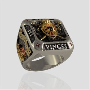 Puede incluir: Un anillo de plata con un dise&ntilde;o en negro y dorado. El anillo presenta un escudo con una cruz y una corona, dos espadas y las palabras "Cross and Crown" y "VINCES".