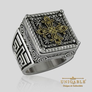Pode incluir: Um anel de prata com uma parte superior quadrada que apresenta um design intrincado. O centro do quadrado é uma cruz dourada com um padrão floral. O anel tem uma banda texturizada com um design geométrico.