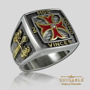 Könnte beinhalten: Ein silberner Ring mit einem roten und goldenen Kreuz und der Inschrift "Hoc Signo Vinces" auf der Band. Das Kreuz hat einen kleinen Diamanten in der Mitte.