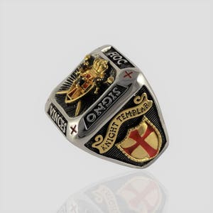 Puede incluir: Un anillo de plata y oro con un dise&ntilde;o de esmalte negro que presenta un escudo de caballero templario con una cruz roja, dos espadas y un casco. El anillo est&aacute; inscrito con las palabras "Hoc Signo Vinces" y "Caballero Templario".
