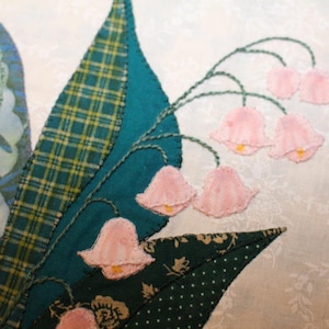 May include: Close-up of a handmade quilt block featuring pink and white lily of the valley flowers with green leaves. The flowers are appliqued and embroidered with a satin stitch.