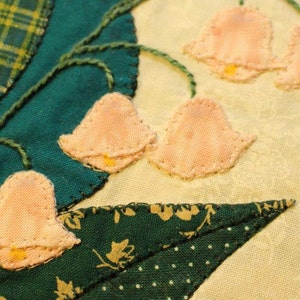 May include: Close-up of a handmade quilt block featuring a cluster of pink lily of the valley flowers with green leaves and stems. The flowers are appliqued and stitched onto a cream-colored background.