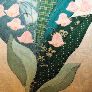 May include: A close-up of a handmade quilt block featuring a cluster of pink lily of the valley flowers with green leaves. The quilt block is made from various shades of green fabric with a patchwork design.