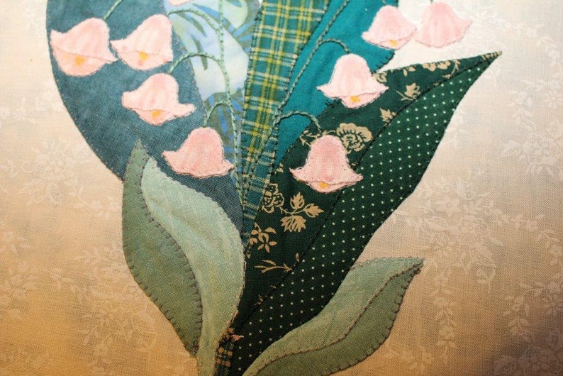 May include: A close-up of a handmade quilt block featuring a cluster of pink lily of the valley flowers with green leaves. The quilt block is made from various shades of green fabric with a patchwork design.