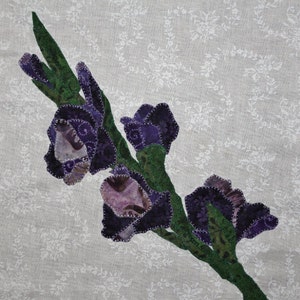 May include: A purple and green fabric flower appliqué on a white background. The flower is made of multiple fabric pieces sewn together to create a three-dimensional effect.