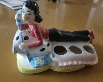 Może przedstawiać: Ceramiczny stojak na szczoteczki do zębów z figurką kobiety w czerwonej koszuli i czarnych spodniach, leżącej na poduszce z niebieskim telefonem. Stojak ma pięć miejsc na szczoteczki do zębów.