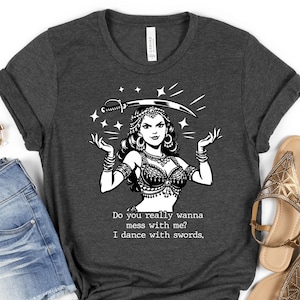 May include: Dark gray t-shirt featuring a black and white illustration of a woman balancing a sword on her head. The text reads, "Do you really wanna mess with me? I dance with swords."
