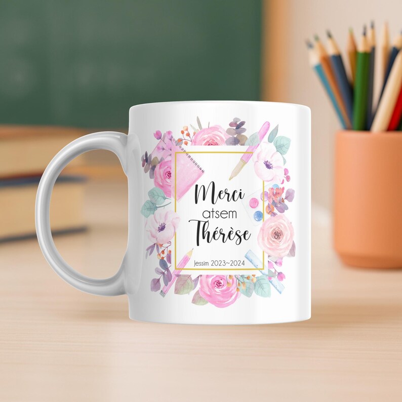 May include: White ceramic mug featuring a floral design with pink roses and watercolor elements. The mug has the text "Merci atsem Th&eacute;r&egrave;se Jessim 2023-2024" in a gold frame. The mug is on a wooden surface.
