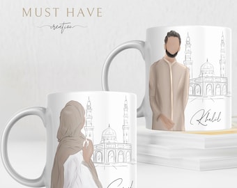 May include: Two white ceramic mugs with illustrations of a mosque and a couple in traditional clothing. The mug on the left has the name "Souad" written on it, and the mug on the right has the name "Khalid" written on it.
