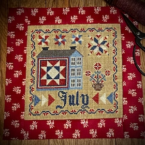 May include: A cross-stitch sampler with a red, white, and blue color scheme. The sampler features a house, a quilt block, flowers, and the word "July".