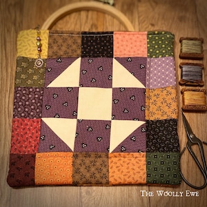 May include: A patchwork quilt bag with a purple and white star design. The bag is made of various fabrics with floral patterns and is lined with a solid purple fabric. The bag is sitting on a wooden surface with a wooden hoop, spools of thread, and scissors.