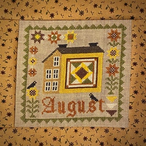 May include: Cross-stitch embroidery of a house with a quilt block, sunflowers, and birds. The text "August" is stitched below the house.