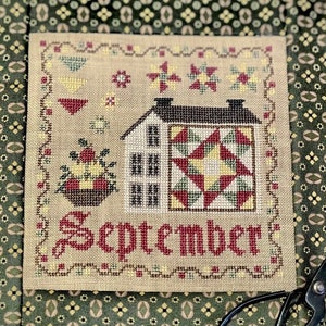 May include: Cross-stitch sampler with a beige background featuring a white house with a red and green quilt block, a basket of red and yellow flowers, and the word "September" in red lettering.