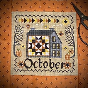 May include: Cross-stitch pattern of a house with a quilt block on the front, two pumpkins, and the word "October". The house is gray with white windows and a black roof. The quilt block is black, white, and yellow. The pumpkins are orange with green stems. The background is a beige fabric with a brown and white pattern.
