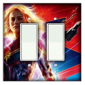May include: A double light switch plate featuring a colourful image of Captain Marvel, a superhero from the Marvel Cinematic Universe. The image shows Captain Marvel in a red, blue, and gold costume, with a glowing energy effect around her. The background is a mix of red, blue, and purple colours.
