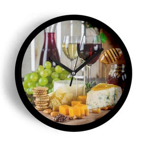 May include: A round wall clock with a black frame. The clock face features a still life of wine, cheese, grapes, and crackers. Wine glasses and bottles are visible in the background. The clock hands point to approximately 10:10.