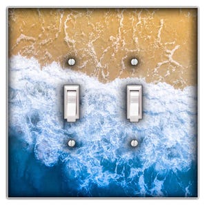May include: A decorative light switch cover featuring an ocean wave design. The image shows a transition from a sandy beach to the blue ocean, with white waves. Two white light switches are in the center, with silver screws.