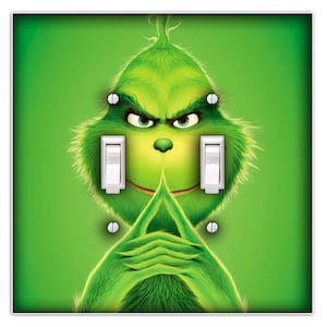 May include: A green light switch cover featuring the Grinch with white light switches and silver screws. The Grinch's face is centered, with his hands clasped below the switches. The word "OFF" is visible on the switches.