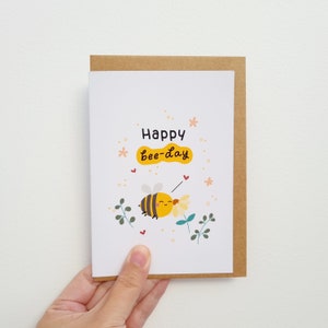 Peut inclure: Une carte blanche avec une enveloppe brune. La carte repr&eacute;sente une abeille de dessin anim&eacute; mignonne avec des rayures jaunes et noires, qui recueille du pollen d'une fleur. La carte dit "Happy Bee-day" en &eacute;criture noire.