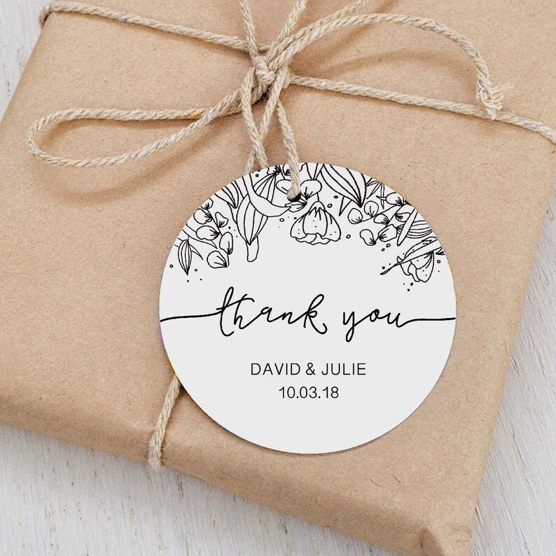 Op de afbeelding: Een ronde witte cadeaulabel met een bloemenontwerp en de woorden "thank you" in zwart script. De label bevat ook de namen "DAVID & JULIE" en de datum "10.03.18". De label is met touw aan een verpakt cadeau bevestigd.