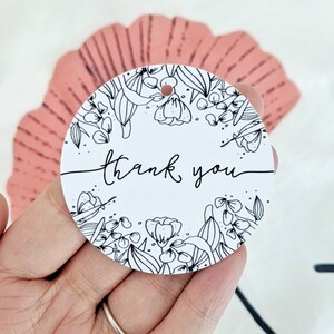 Op de afbeelding: Een witte, ronde cadeaulabel met de woorden "thank you" in cursief schrift. De label is omcirkeld door een zwart-wit bloemenontwerp. Er zit een klein gaatje aan de bovenkant om te rijgen. De label wordt in een hand gehouden.