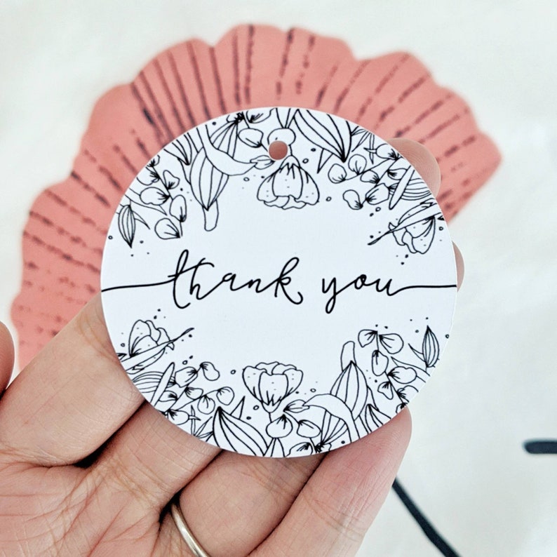 Op de afbeelding: Een witte, ronde cadeaulabel met de woorden "thank you" in cursief schrift. De label is omcirkeld door een zwart-wit bloemenontwerp. Er zit een klein gaatje aan de bovenkant om te rijgen. De label wordt in een hand gehouden.
