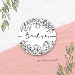 Op de afbeelding: Een ronde witte sticker met een bloemenrand en de woorden "thank you" in zwart script. De sticker is omgeven door een aquarelontwerp van groene bladeren en een roze achtergrond.