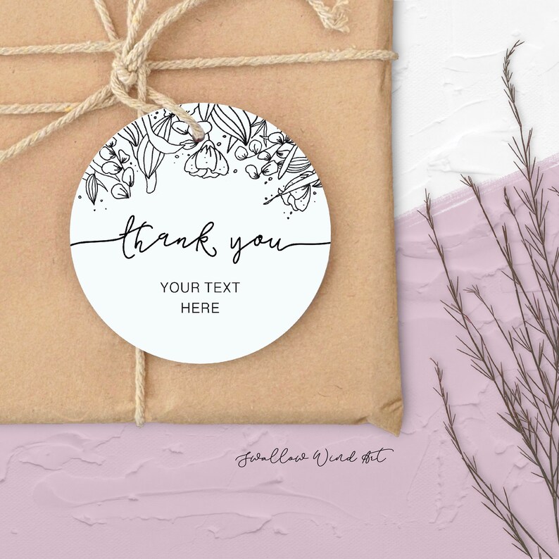 Op de afbeelding: Een ronde witte cadeaulabel met een bloemenontwerp en de woorden "thank you" in zwart script. De tag bevat ook de tekst "YOUR TEXT HERE". De tag is met touw aan een bruin pakket bevestigd.