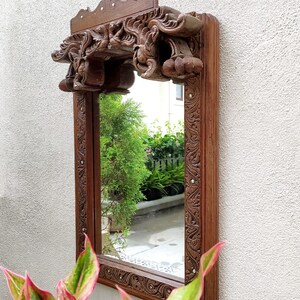 May include: A rectangular, wooden mirror with intricate carvings. The frame and decorative top are a rich brown color. The mirror is mounted on a white wall, with greenery visible in the reflection and foreground.