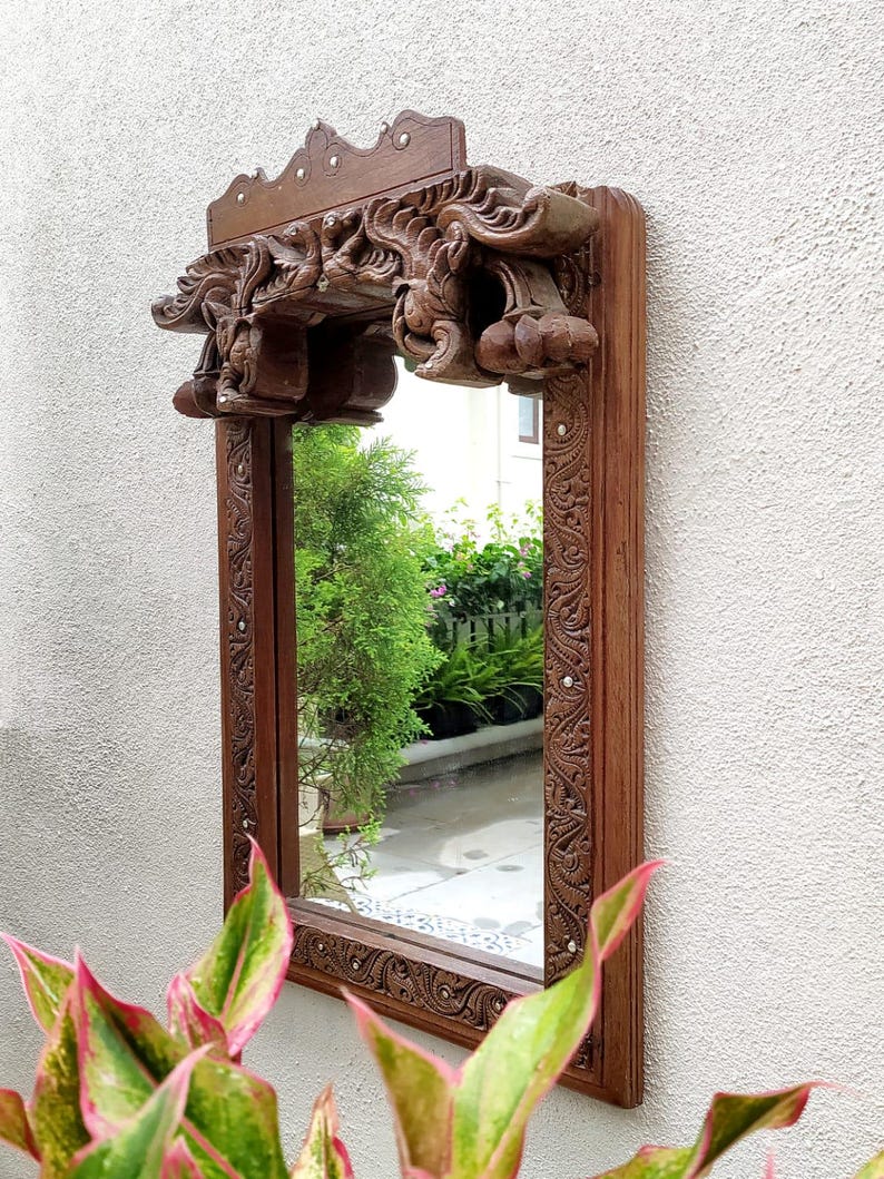 May include: A rectangular, wooden mirror with intricate carvings. The frame and decorative top are a rich brown color. The mirror is mounted on a white wall, with greenery visible in the reflection and foreground.