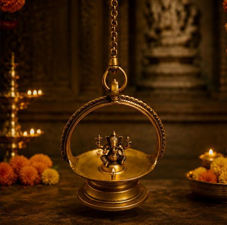 May include: A hanging brass oil lamp featuring a central figure of Ganesha seated within a circular frame. The lamp is suspended by a metal chain and includes a small lit wick at the base of the deity, casting a warm glow on the metallic surface.