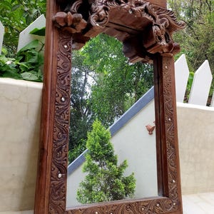 May include: A rectangular, ornate wooden mirror with a carved frame. The frame is dark brown with intricate floral and scrollwork designs, and small silver studs. The mirror reflects a green tree and the sky.