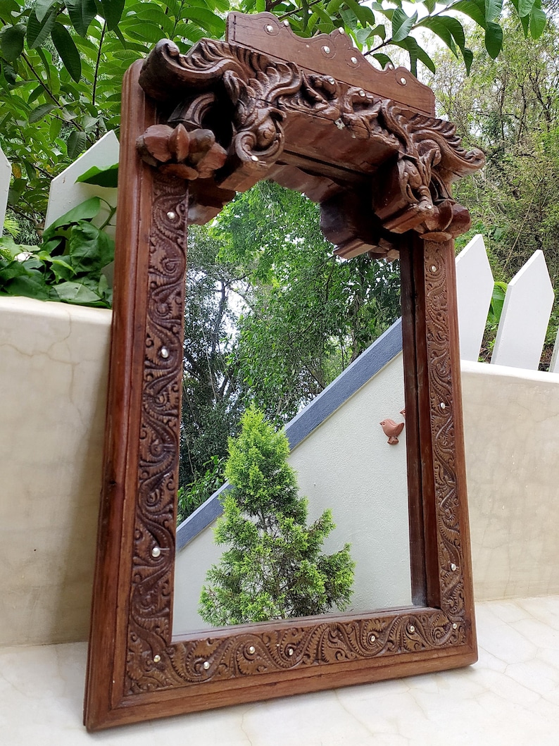 May include: A rectangular, ornate wooden mirror with a carved frame. The frame is dark brown with intricate floral and scrollwork designs, and small silver studs. The mirror reflects a green tree and the sky.