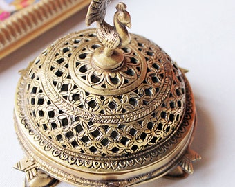 May include: A gold metal incense burner with a peacock design on the lid. The burner has a round base with four legs and a decorative, openwork design.