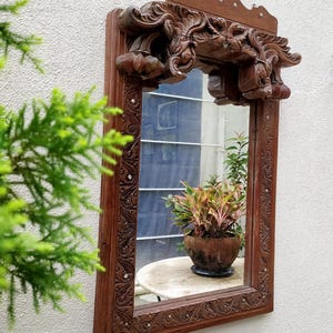 May include: A rectangular, ornate wooden mirror with intricate carvings and a decorative top. The mirror reflects a small table with a potted plant. The frame is a dark brown color, and the carvings add a vintage aesthetic.