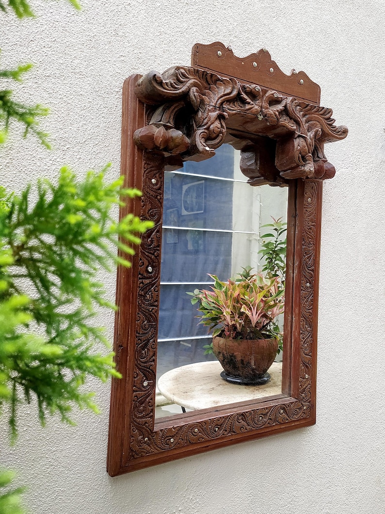 May include: A rectangular, ornate wooden mirror with intricate carvings and a decorative top. The mirror reflects a small table with a potted plant. The frame is a dark brown color, and the carvings add a vintage aesthetic.