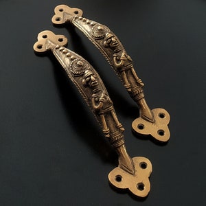 May include: Two ornate brass door handles with intricate designs featuring figures and geometric patterns. The handles are shaped like a curved arch with a decorative end piece.