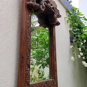 May include: A rectangular, ornate wooden mirror with a dark brown finish. The frame features intricate carvings and a decorative top. The mirror is mounted on a textured, off-white wall, reflecting greenery.